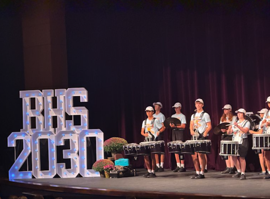 Illuminated marquee letters reading "BHS 2030" beside a student drumline performing in matching white shirts and caps on a decorated stage.