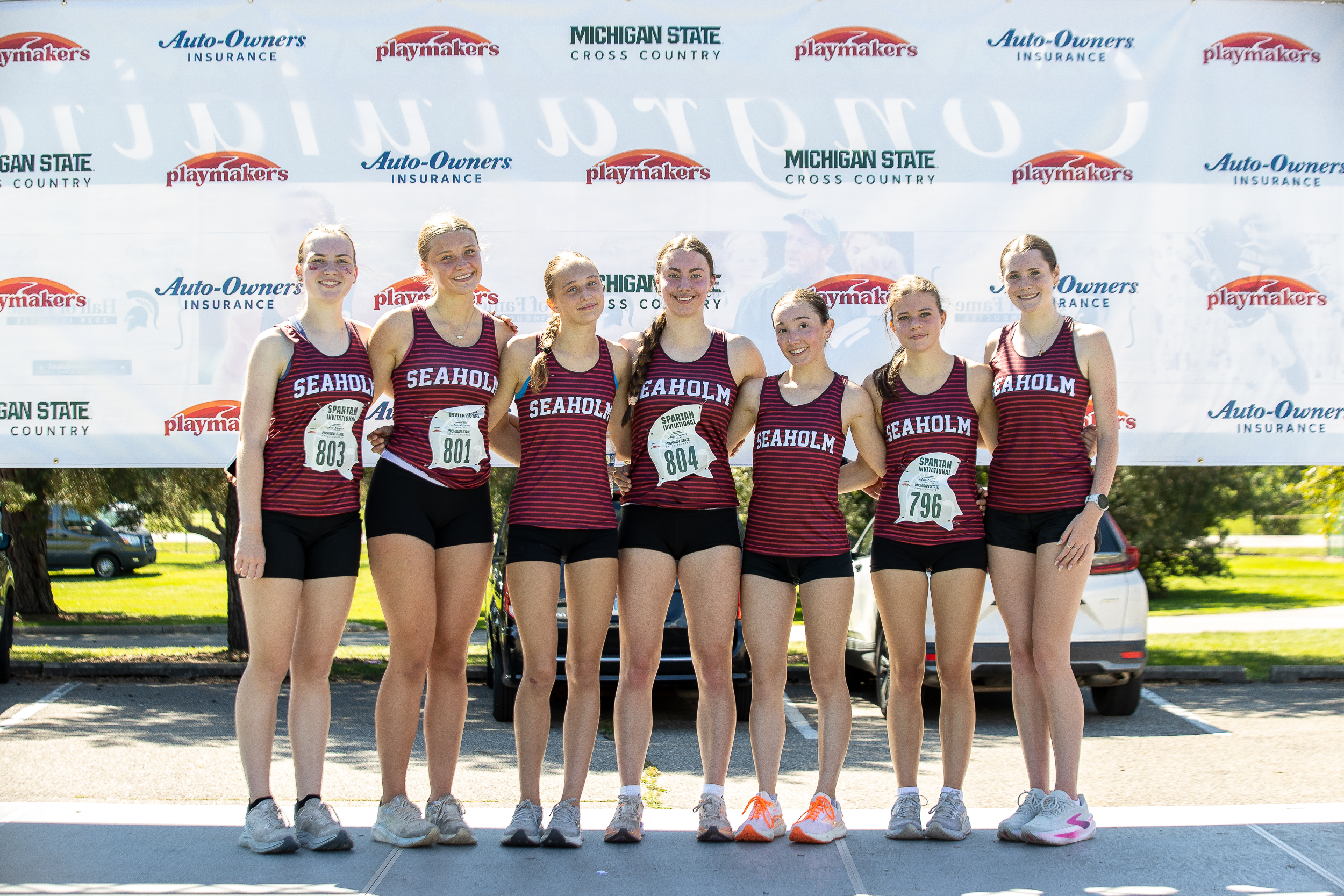 Seven Birmingham Seaholm girls cross country runners in maroon-and-black striped Seaholm singlets stand arm-in-arm in front of a sponsor banner featuring Auto-Owners Insurance, Playmakers and Michigan State Cross Country logos.