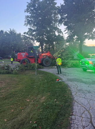 A DPW crew member in a high-visibility vest works near a Deere loader clearing fallen tree branches from a residential roadway at dusk, with a police vehicle's lights visible in the background