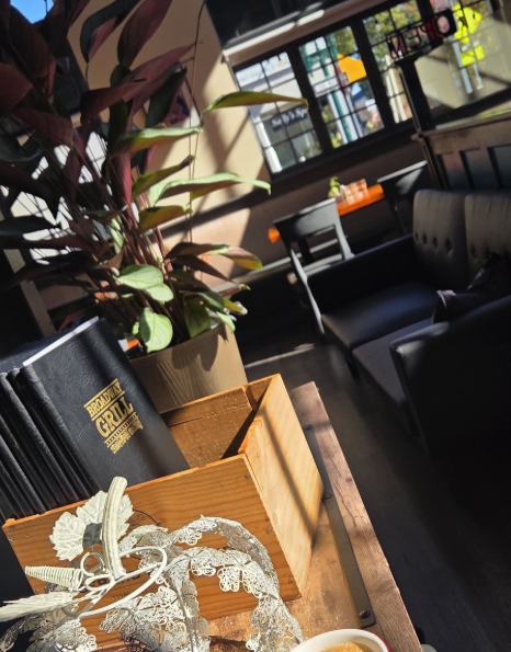 Broadway Grill’s interior with black seating, tables, a potted plant and menus beside the entrance.