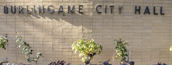 The words 'Burlingame City Hall' in metal lettering on a tan brick building facade, with small potted trees planted along the wall.