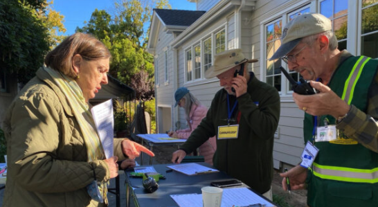 Volunteers at an outdoor table use handheld radios during a simulated disaster drill, with one wearing a badge labeled "Communications" and another in a reflective safety vest.