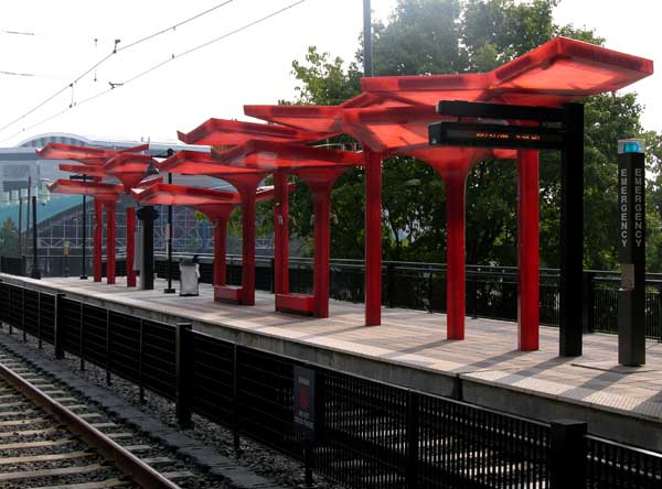 Outdoor light rail platform with red canopy shelters and no fare gates visible.