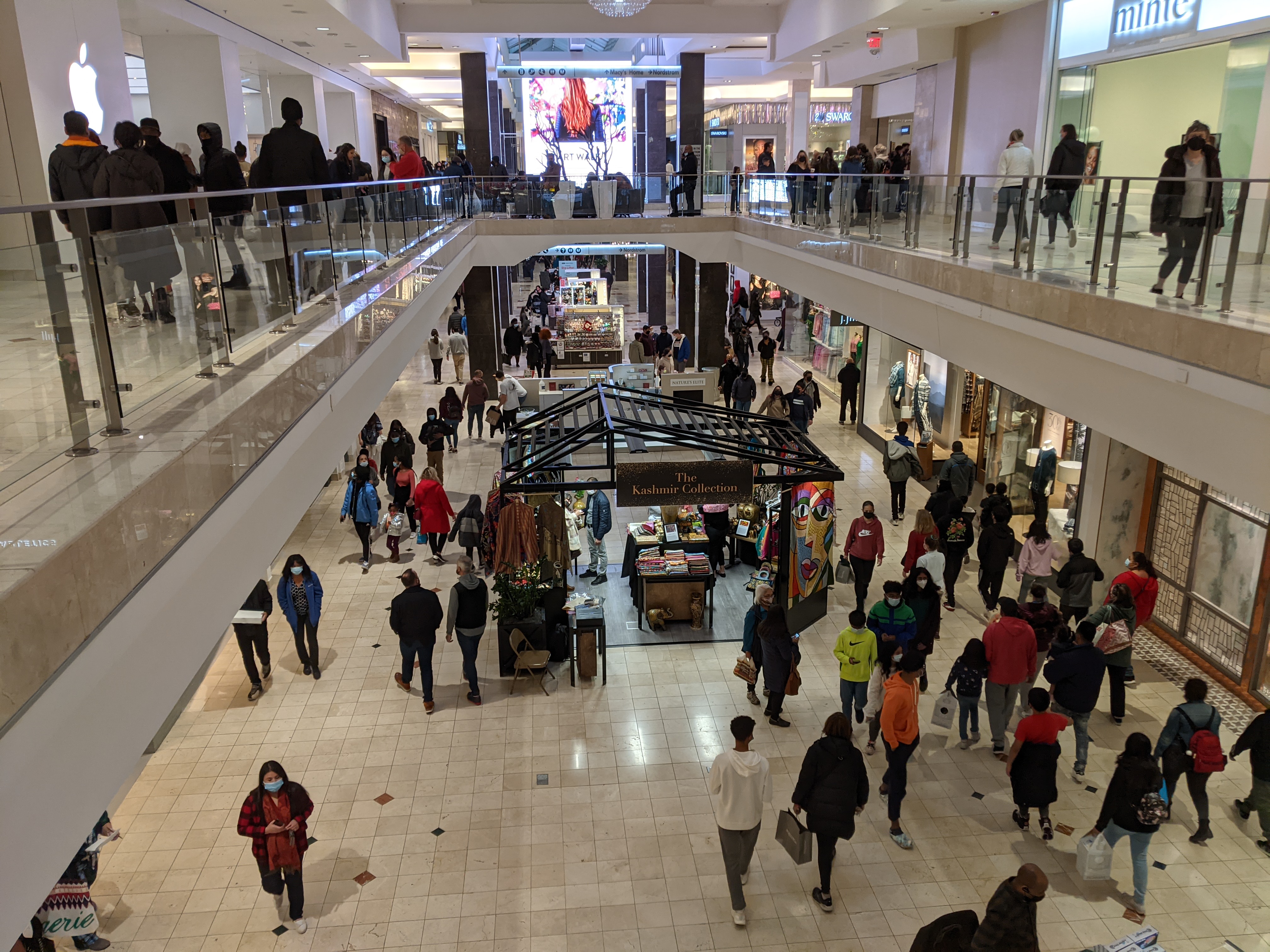 Two-level shopping mall interior with shoppers on both floors and retail storefronts.