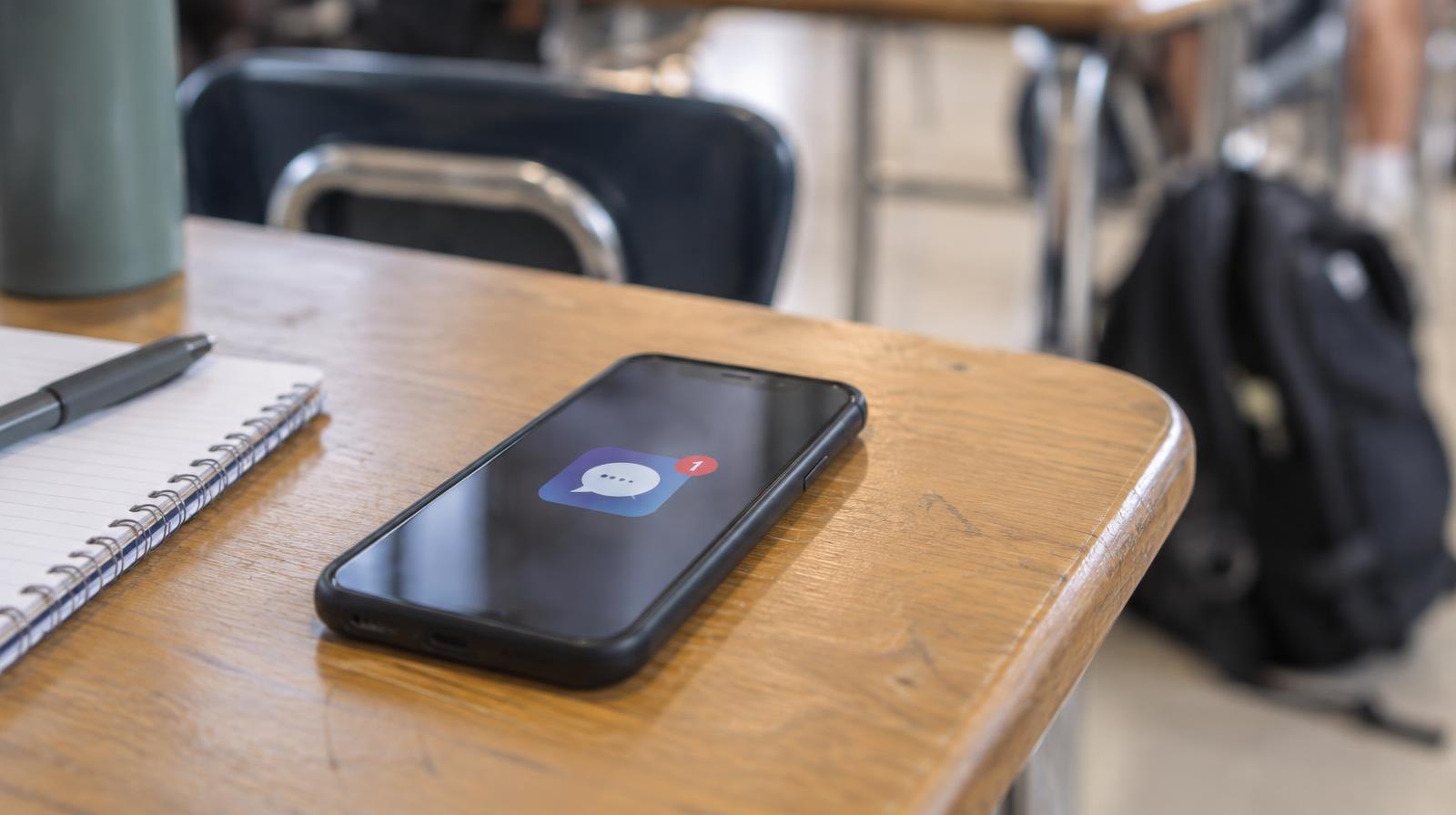 A smartphone face-up on a wooden school desk in a sunlit classroom.