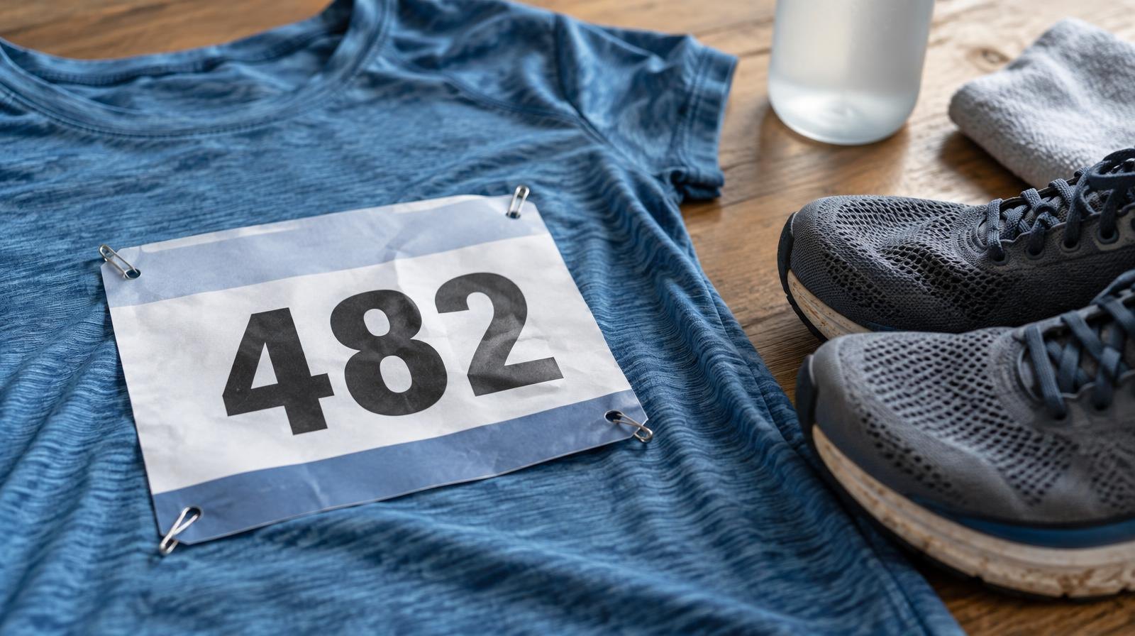 A race bib pinned to a running shirt lying next to a pair of running shoes.