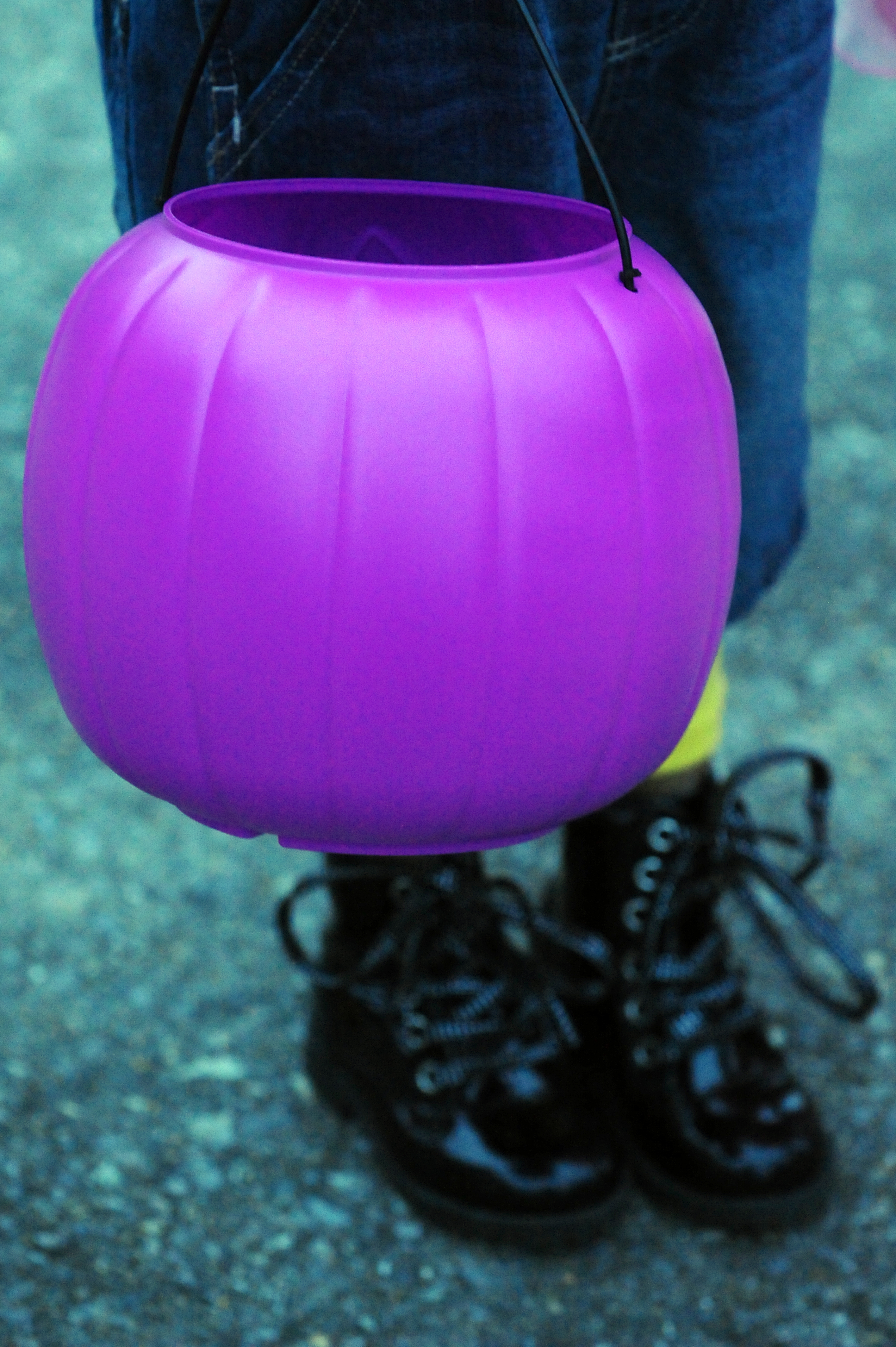 A child in jeans and boots holds a purple plastic pumpkin-shaped candy bucket.