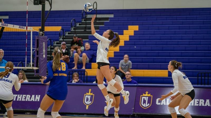 A volleyball player in white jumps to spike at the net in a gymnasium with Cal Lutheran branding.