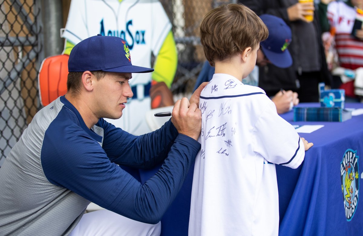Baseball player in AquaSox cap signs the back of a young fan's jersey.