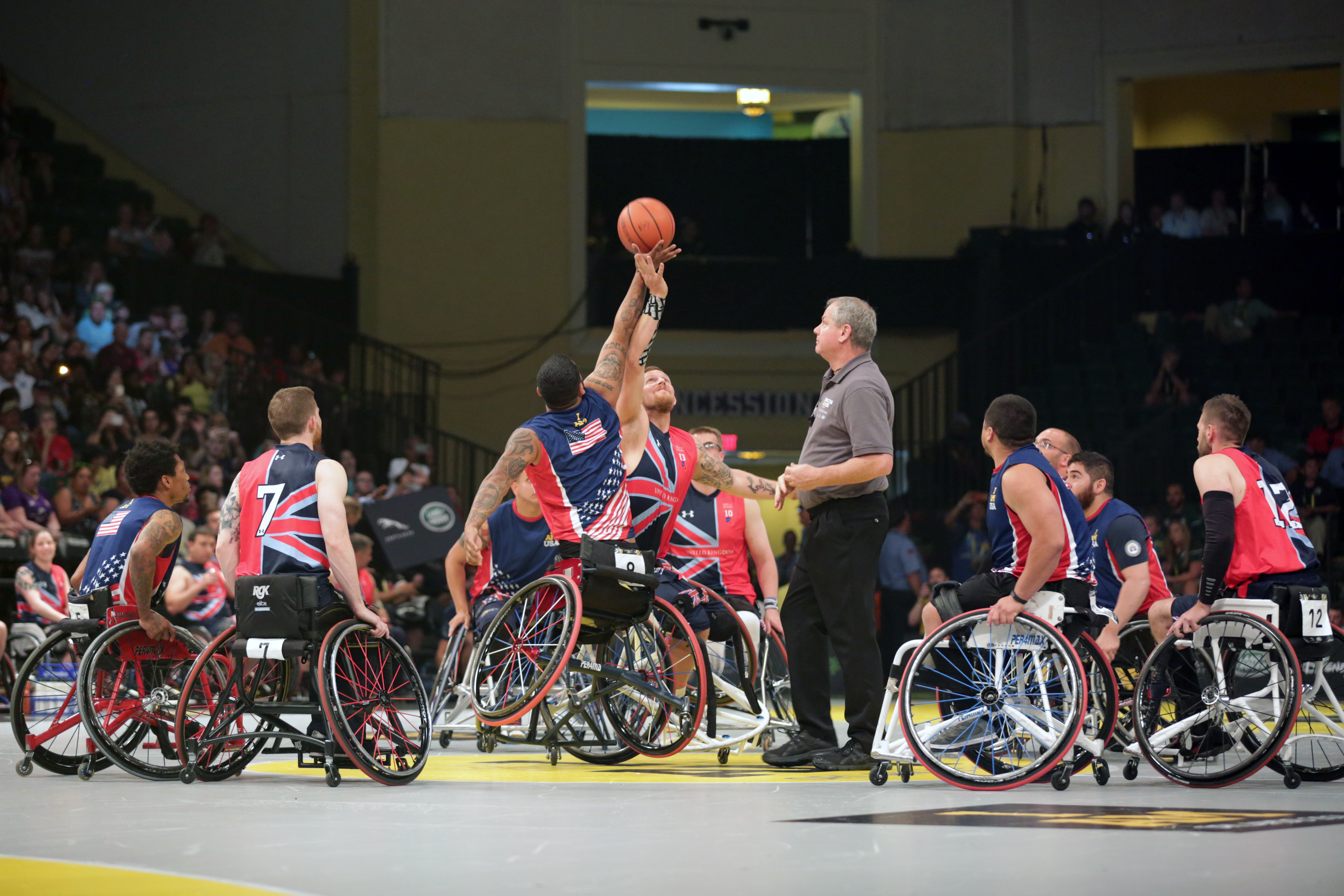 Athletes in wheelchairs compete at a basketball tip-off on an indoor court.