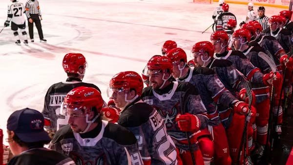 Hockey players in red helmets and camouflage-style jerseys lined up along the team bench.