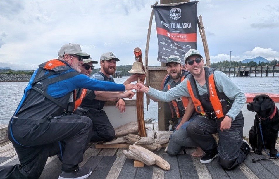 Four sailors in life jackets fist-bump on a dock with a Race to Alaska banner and mountains behind them.