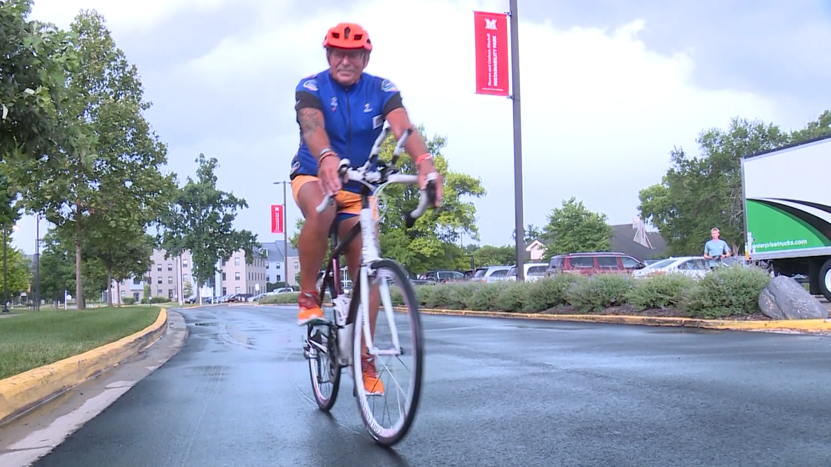 A cyclist in a blue jersey and orange helmet rides a white bicycle on a wet campus road.