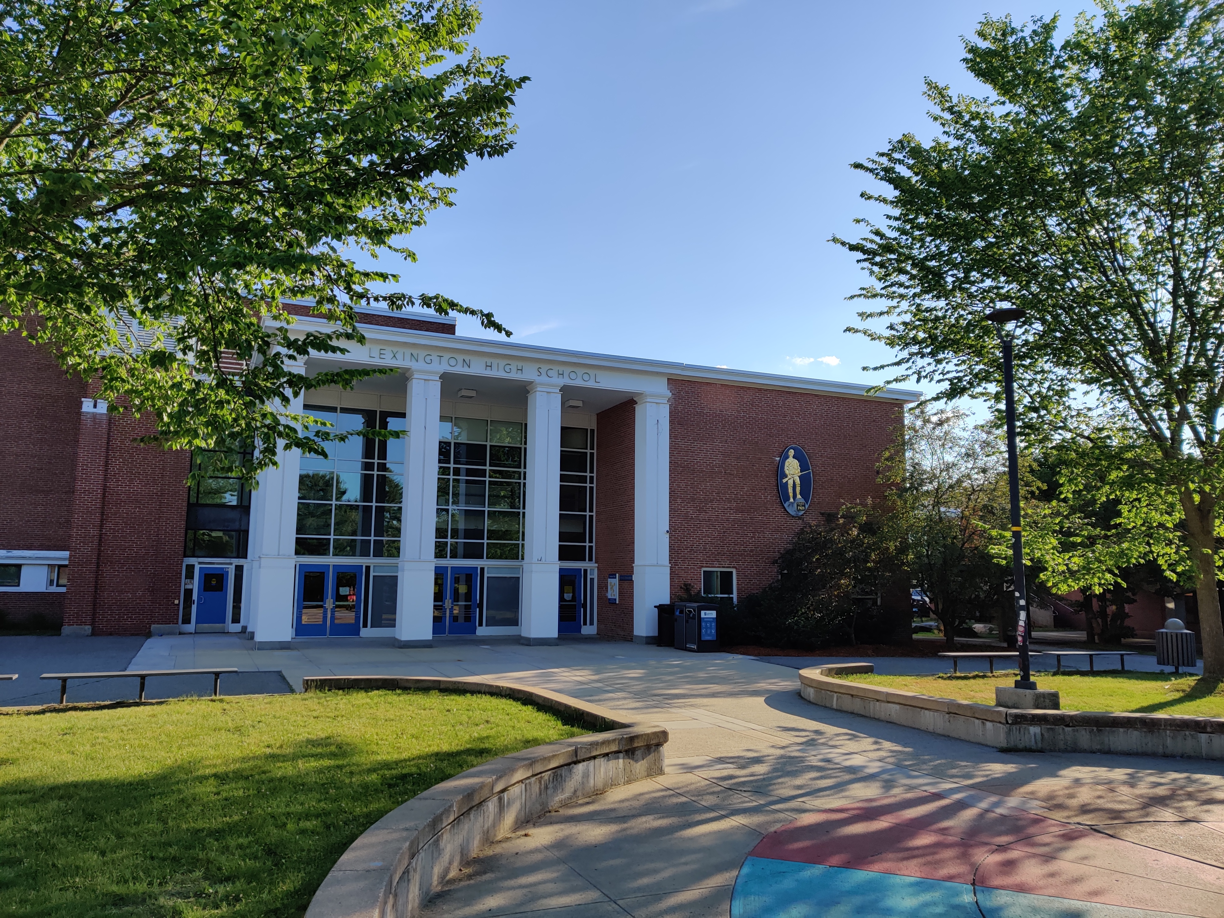 Brick school exterior with white columns, blue doors, and a school emblem, surrounded by trees.