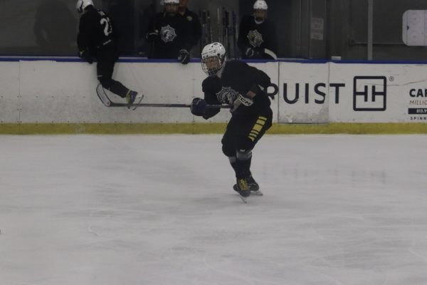 Hockey player in dark uniform and white helmet crouches in a skating stance on indoor ice.