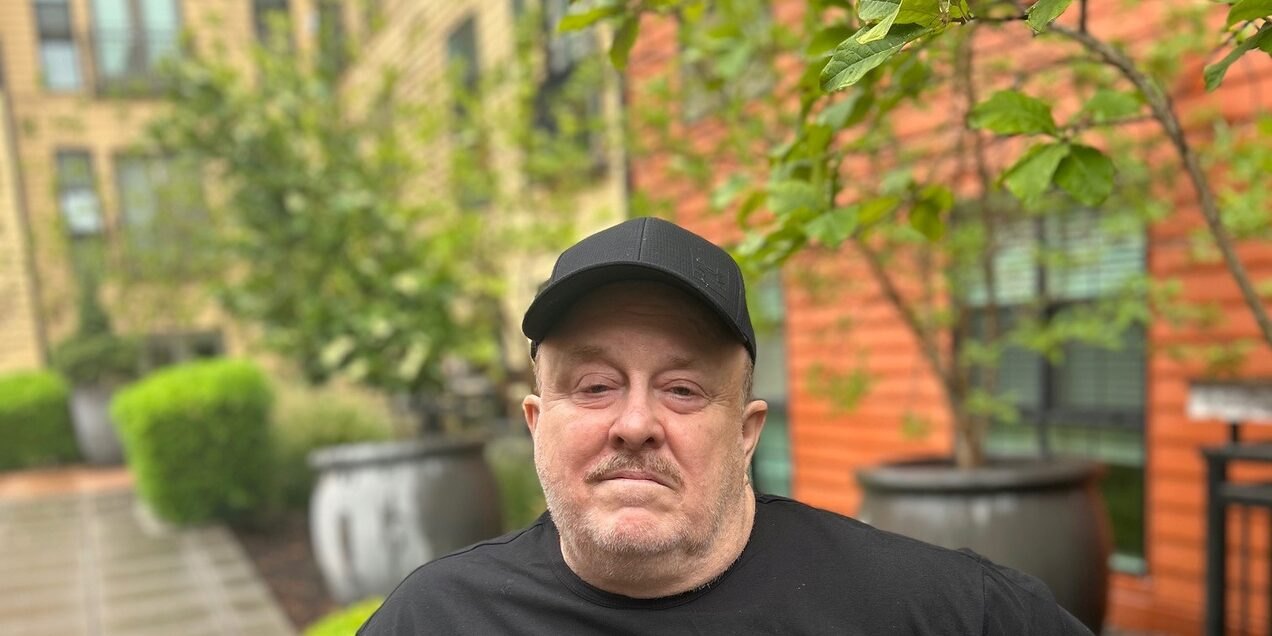 Man in black cap and t-shirt stands outside a brick building with potted trees.