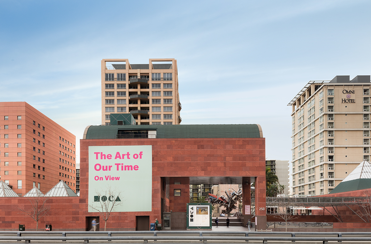 Red sandstone museum building with a banner on its facade, surrounded by urban high-rises.