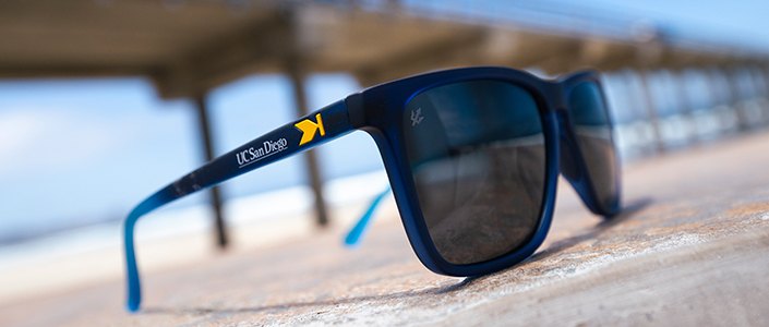 Navy blue sunglasses with UC San Diego branding on the temple arm, resting on a wooden surface with an ocean pier in the background.