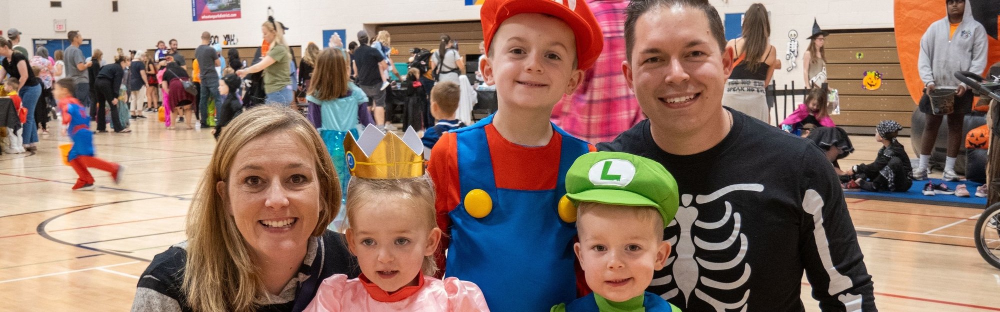 A family of five in Halloween costumes poses inside a gymnasium with costumed attendees in the background.
