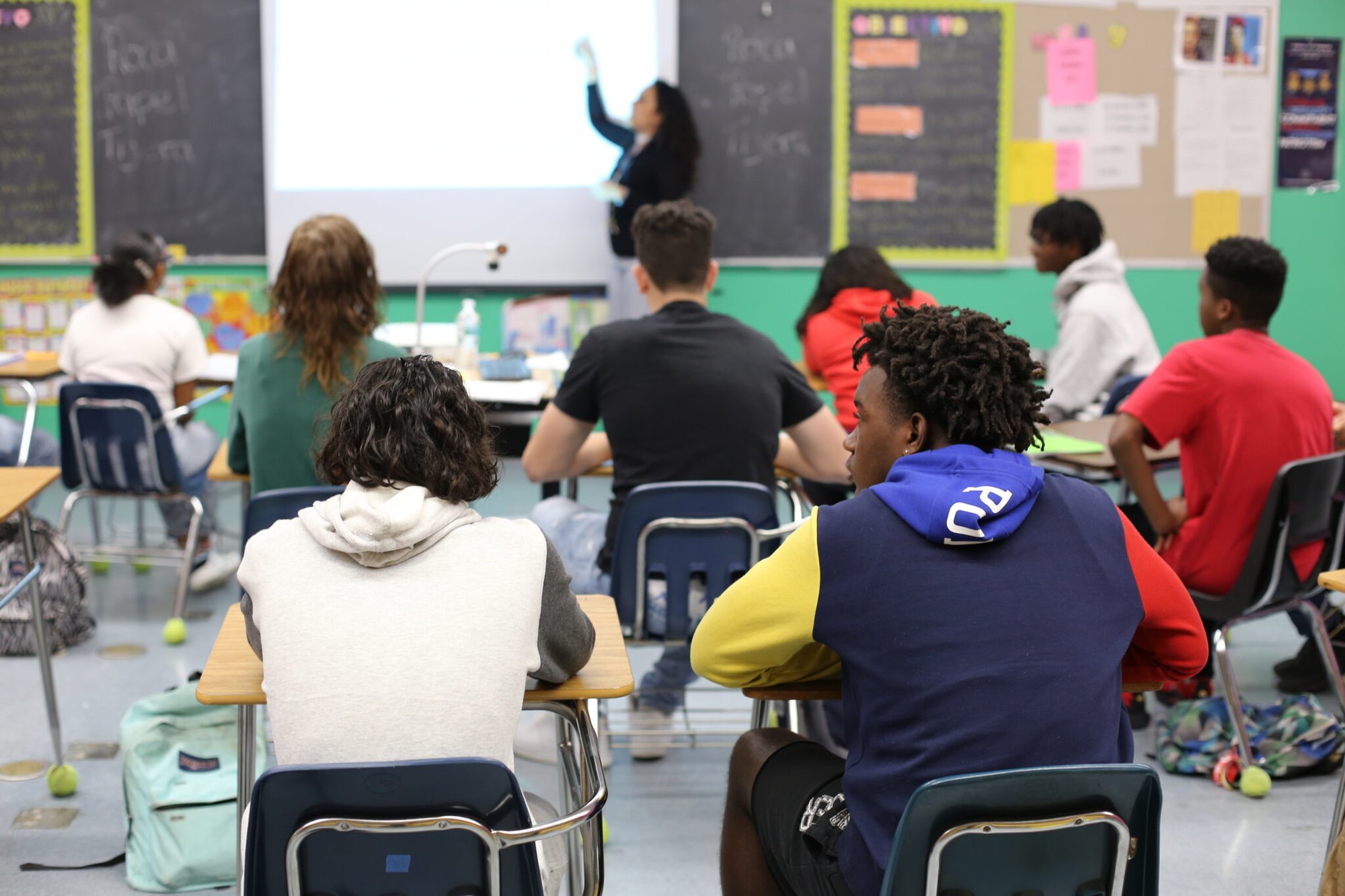 Students seated at desks in a classroom while a teacher points at a projection screen.