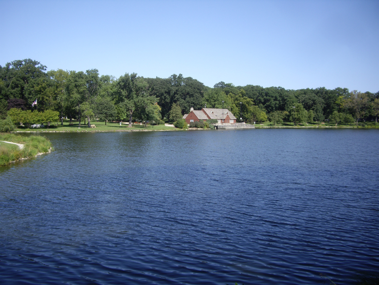 Calm lake with tree-lined shores and a red brick building in the background.