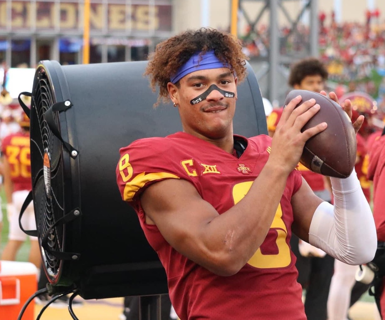 Football player in Iowa State red jersey holding a ball on the sideline.