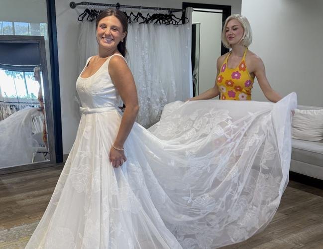 A woman in a white lace wedding gown stands in a bridal shop while another woman holds out the skirt.