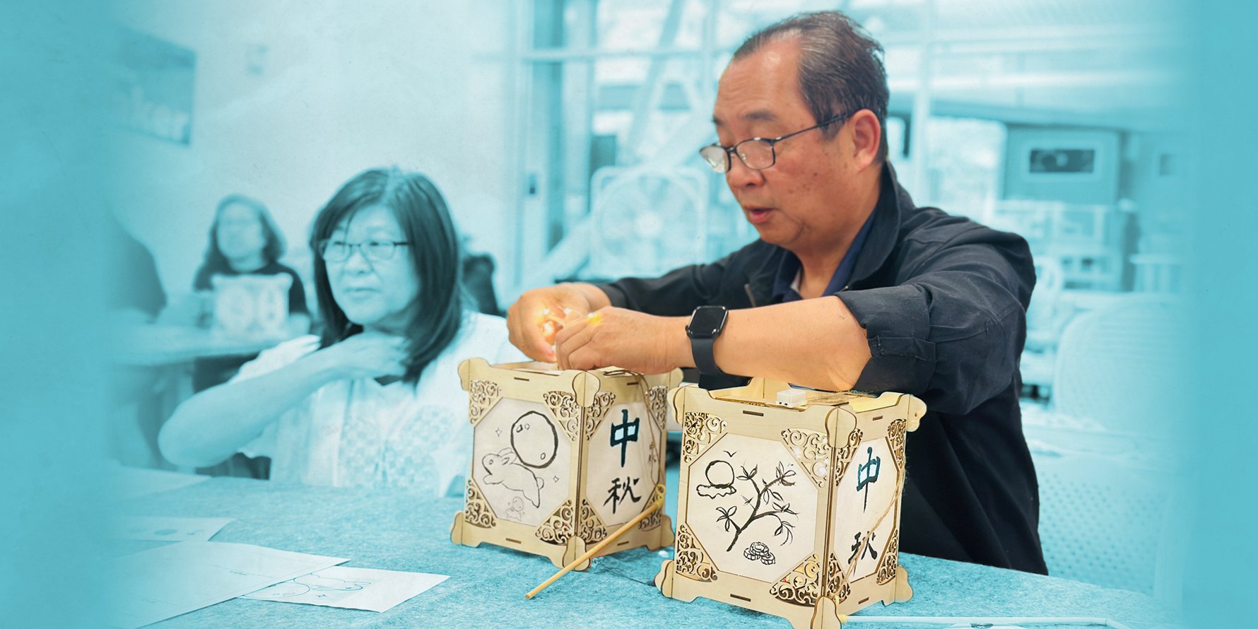 Man in black shirt assembling a decorated square paper lantern at an indoor workshop.