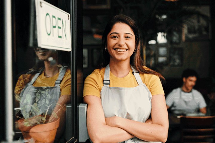 A smiling woman in an apron stands with arms crossed in front of a shop's open door.