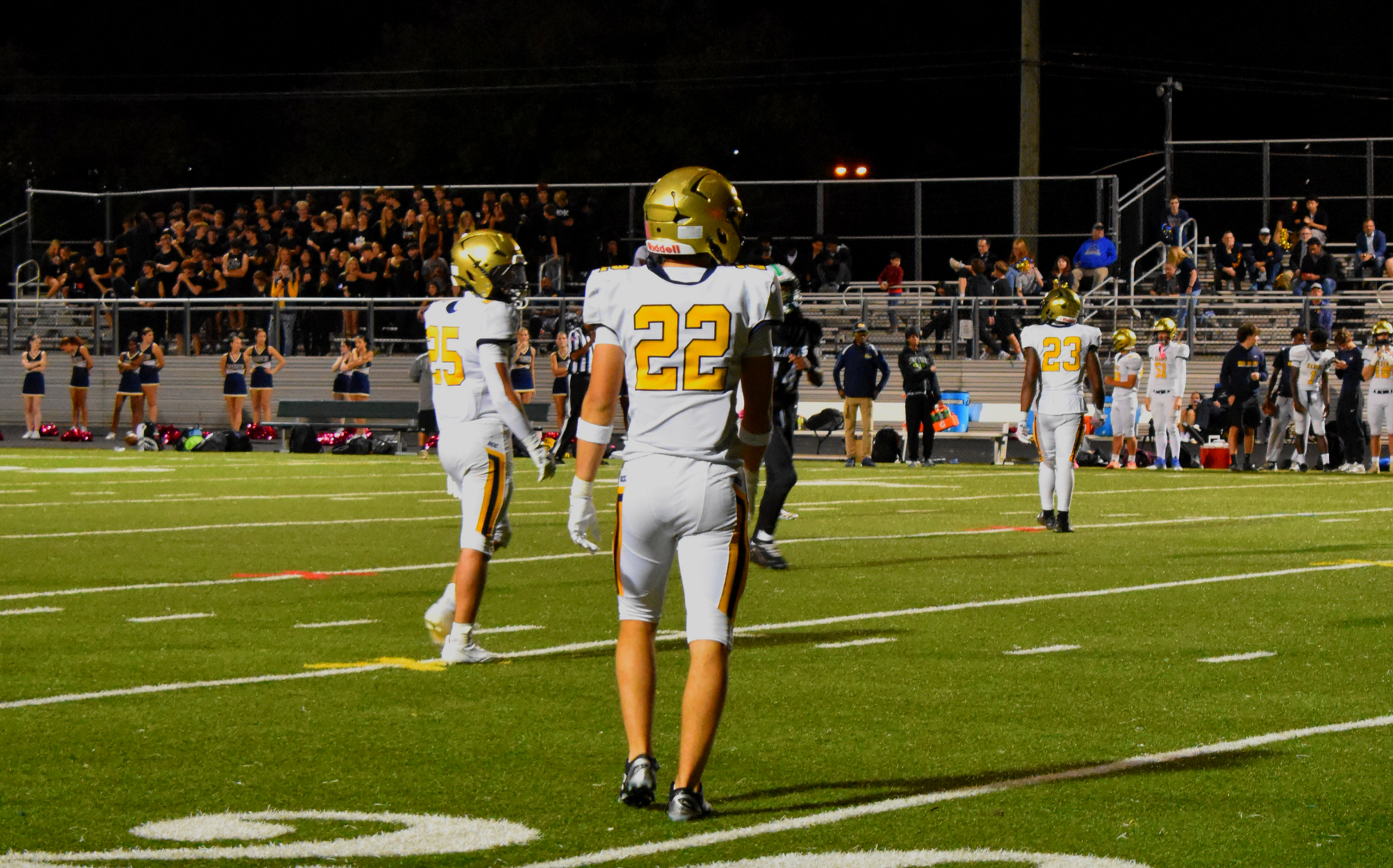 Three football players in white uniforms and gold helmets on a lit field at night with a packed stadium behind them.