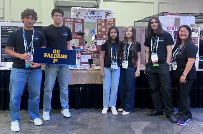 Seven students in matching black shirts stand before a Falcon Drone display booth.