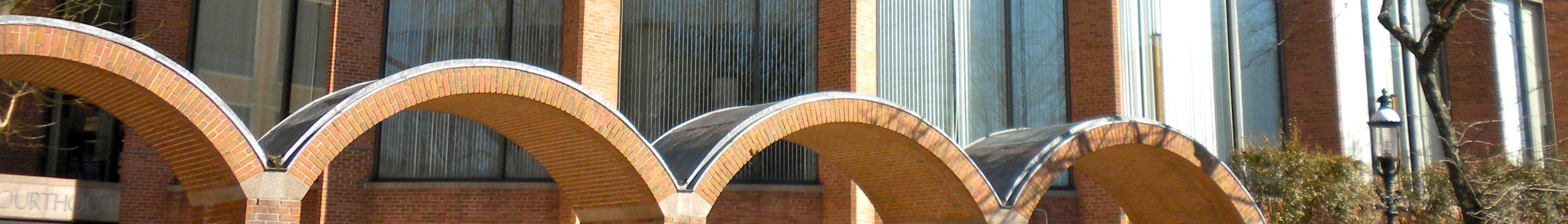 Wide exterior view of a brick courthouse with a row of arched glass panels and a bare tree.