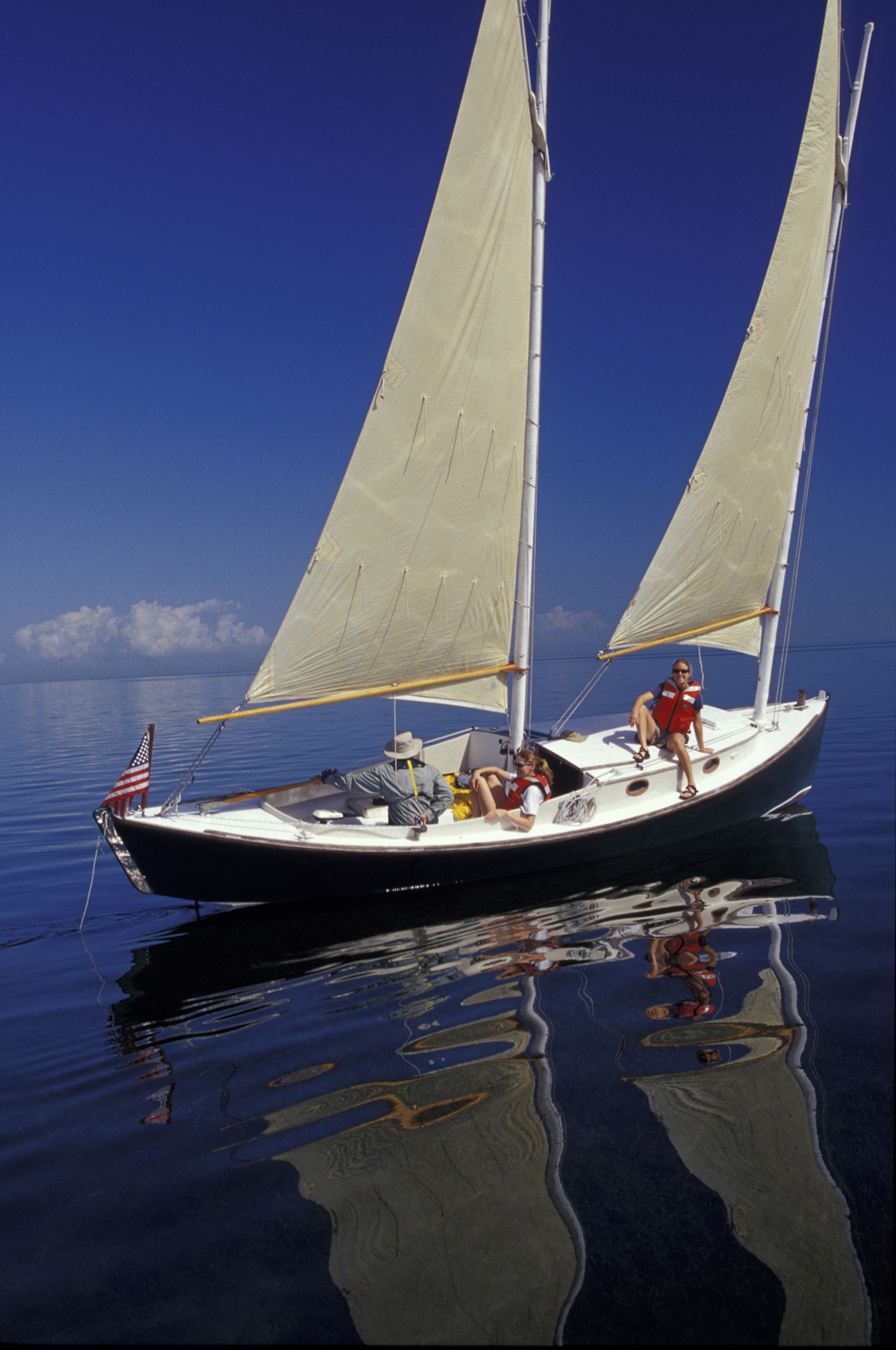A white sailboat with two sails raised moves across still, reflective bay water.