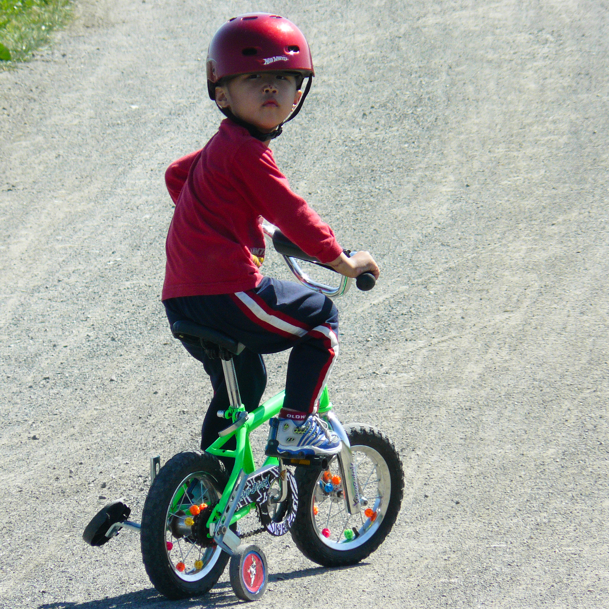 Young child in a red helmet riding a small green bicycle with training wheels on gravel.