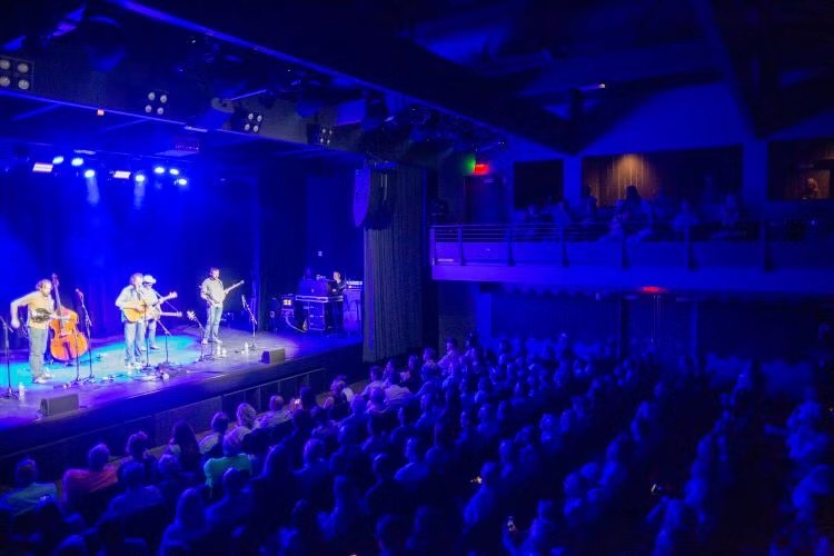 Band performing on a blue-lit stage before a large seated audience in a concert hall.