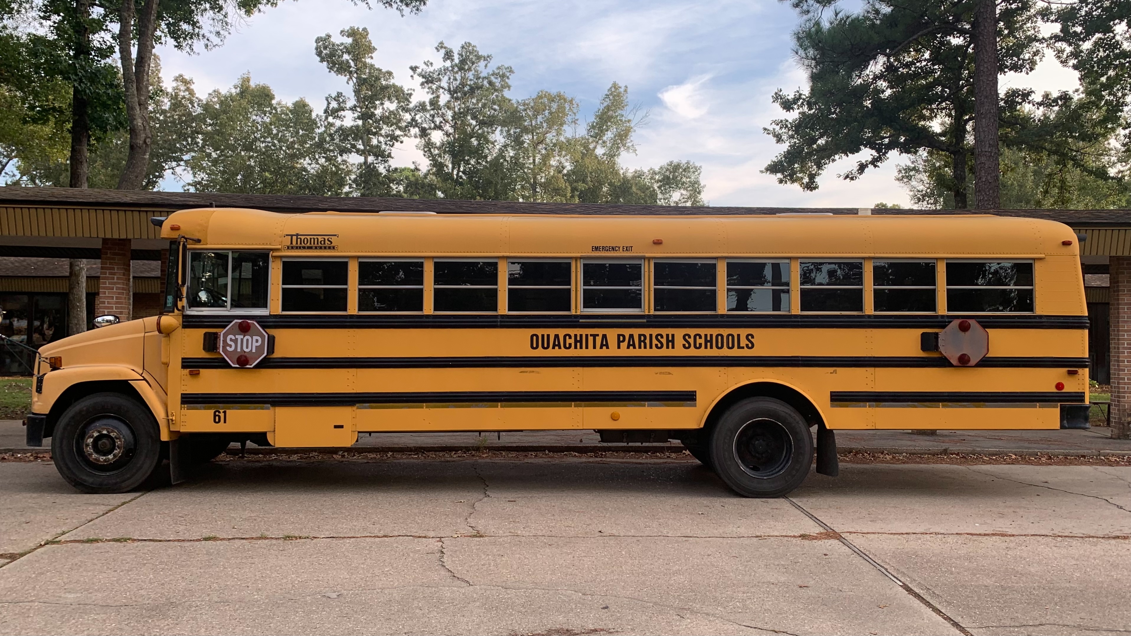 A yellow school bus parked in a lot near a brick building and trees.