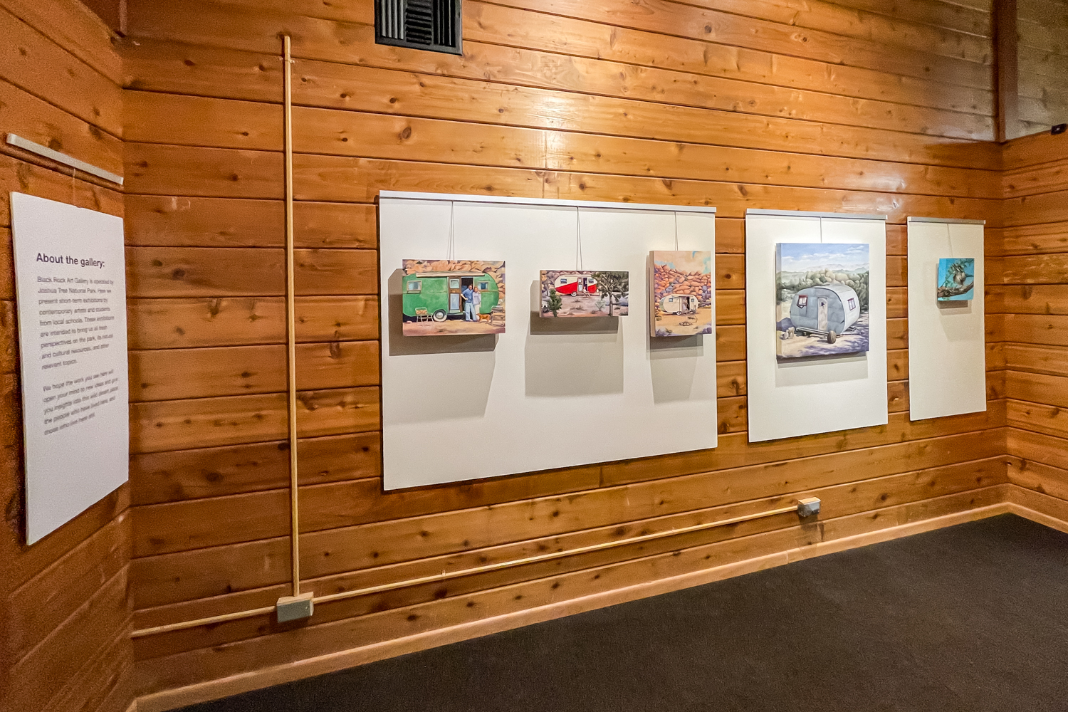 Small colorful paintings mounted on white panels along wood-paneled gallery walls.