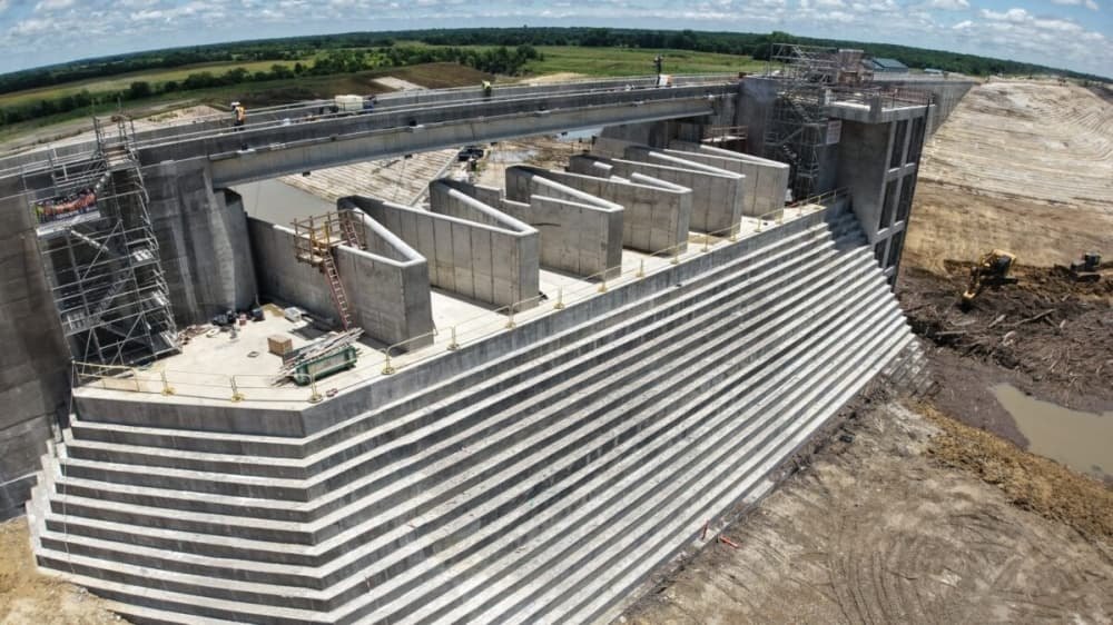 Aerial view of a large concrete dam intake structure under construction with stepped walls and gate bays.
