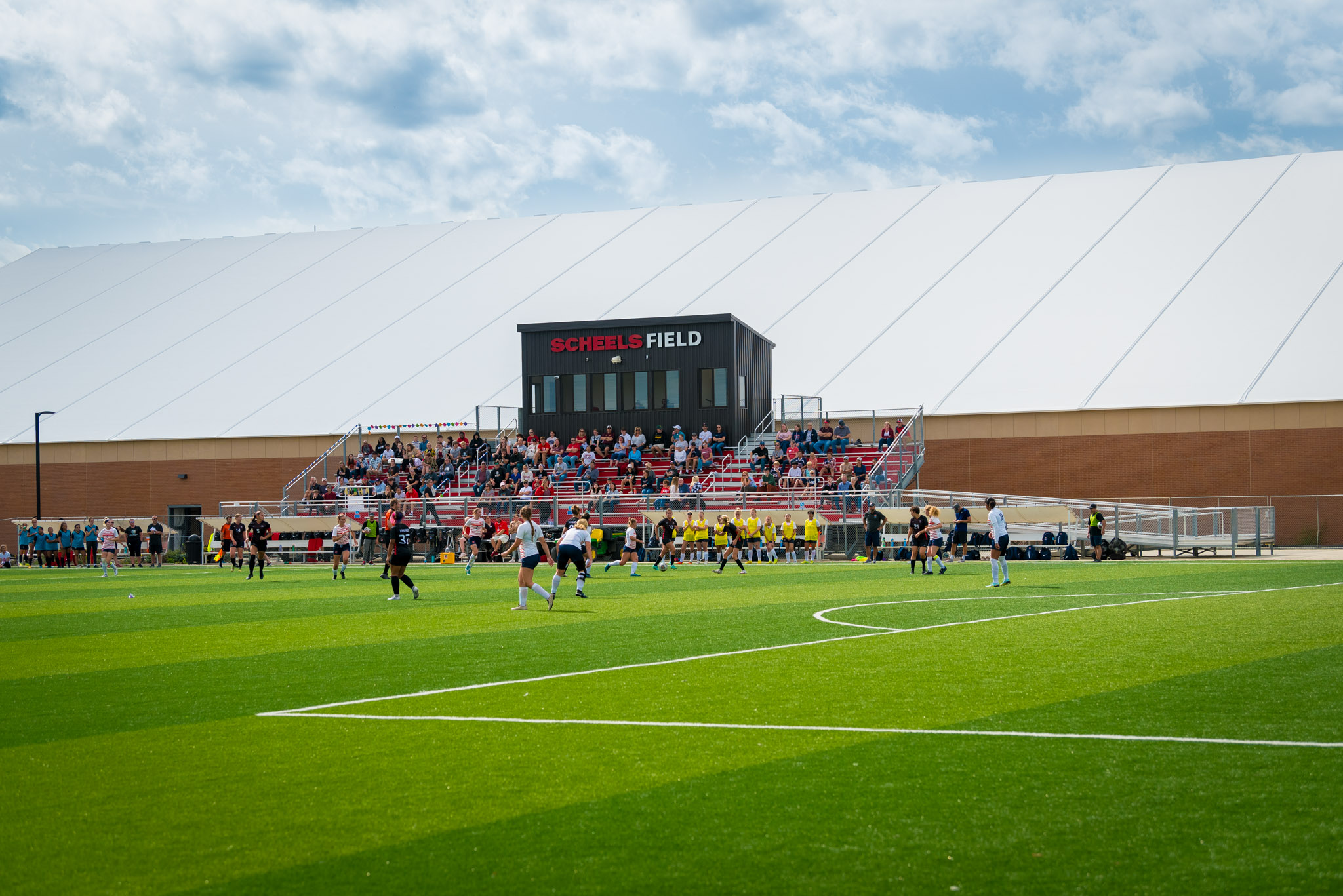 Players compete in a soccer match on a green artificial turf field with spectators in the stands.