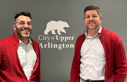 Two men in matching red cardigans smile in front of a City of Upper Arlington wall logo.