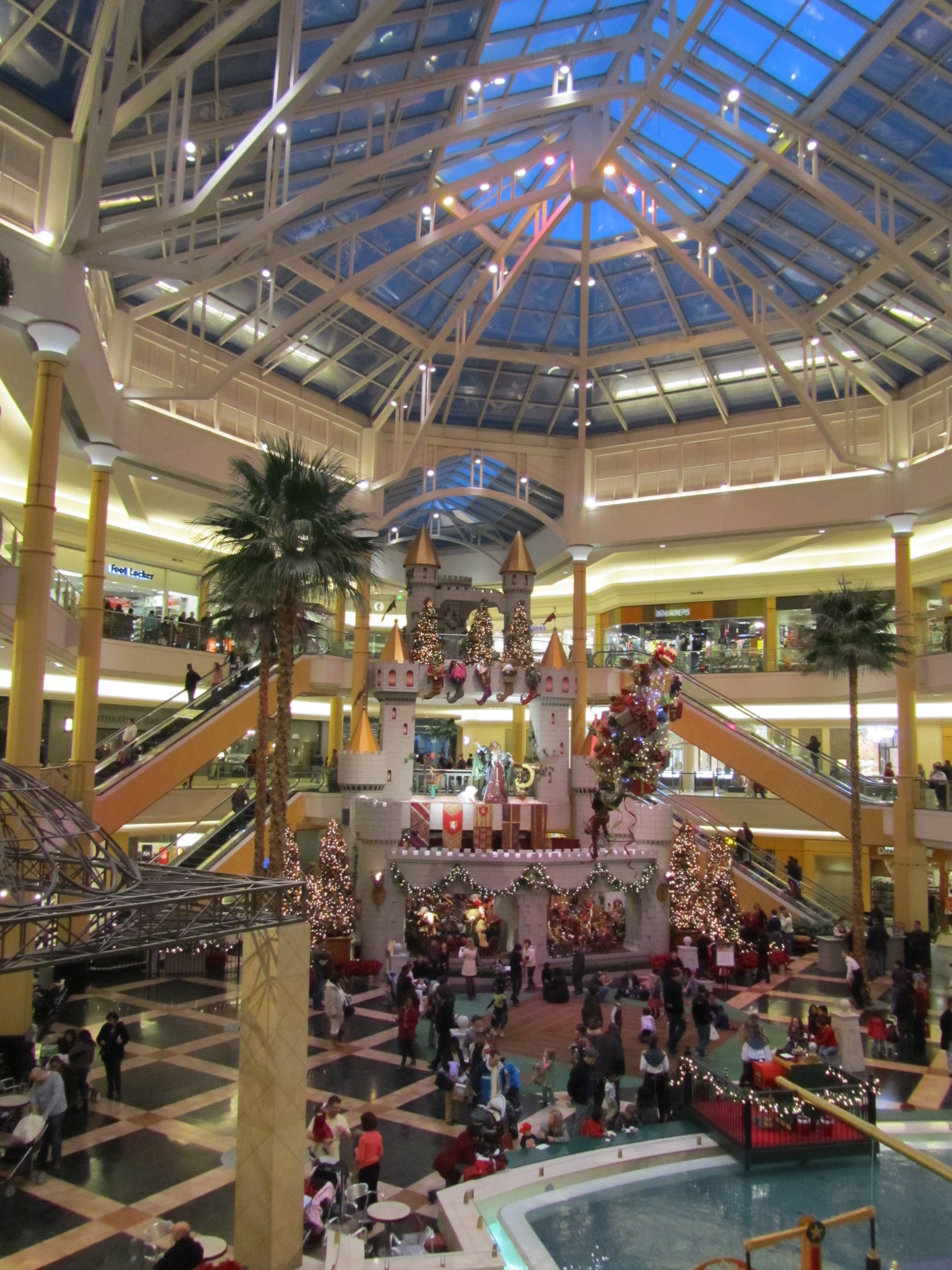 Multi-level mall interior with glass skylight atrium, escalators, and palm trees.