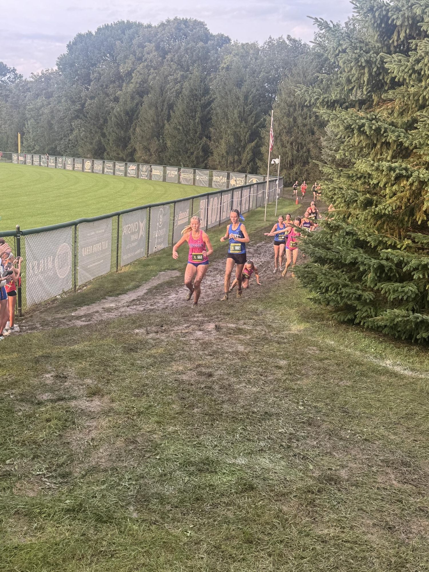 Female cross-country runners race along a muddy dirt path lined with a green banner fence.