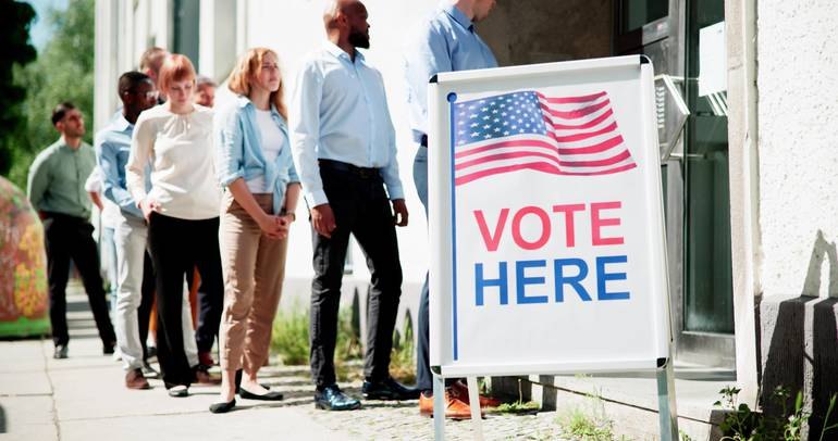 A diverse group of people standing in line outside a building with a Vote Here sign.