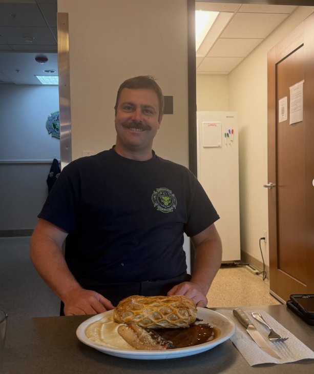 Smiling man in a dark uniform shirt standing behind a plate of food in a firehouse dining area.