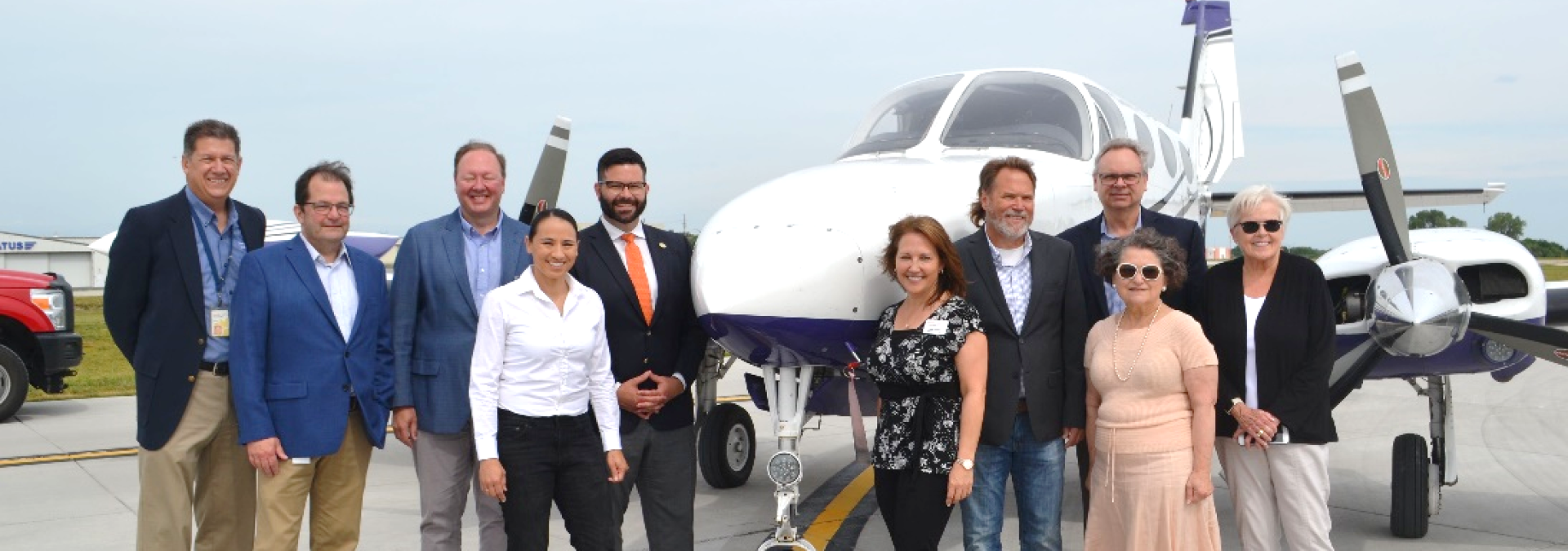 Group of officials standing in front of a small twin-engine aircraft on an airport tarmac.