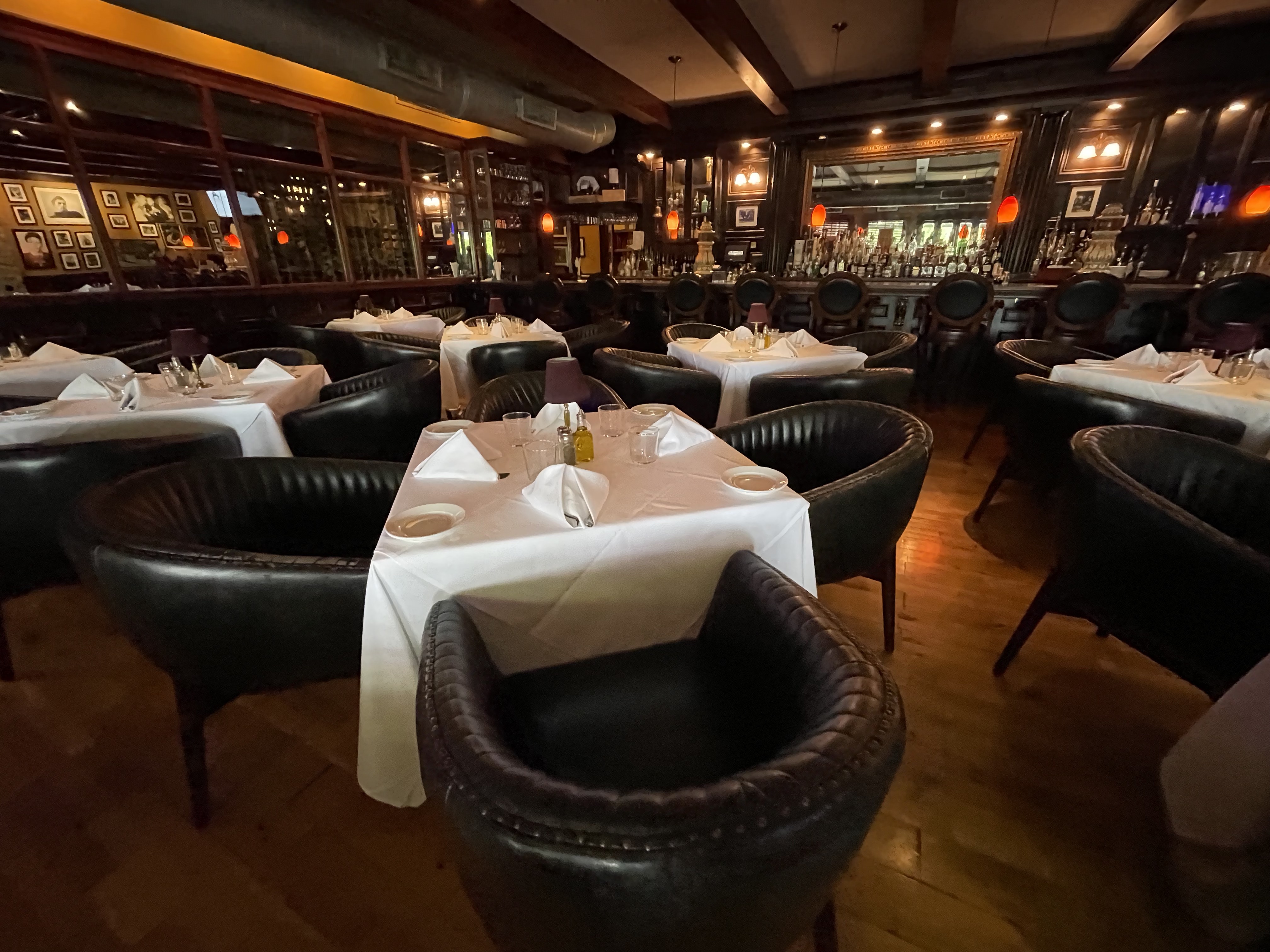 Upscale restaurant dining room with white tablecloths, leather chairs, and a bar in the background.