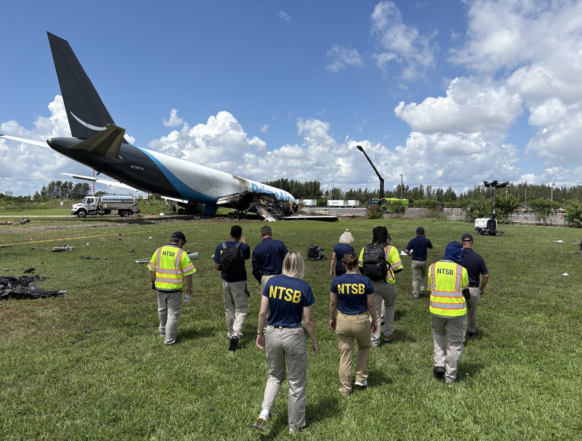 NTSB investigators in yellow vests walk toward a damaged cargo jet resting on grass beyond a runway.