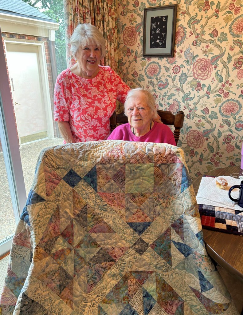 Two older women in pink tops hold up a large patchwork quilt in a floral-wallpapered room.