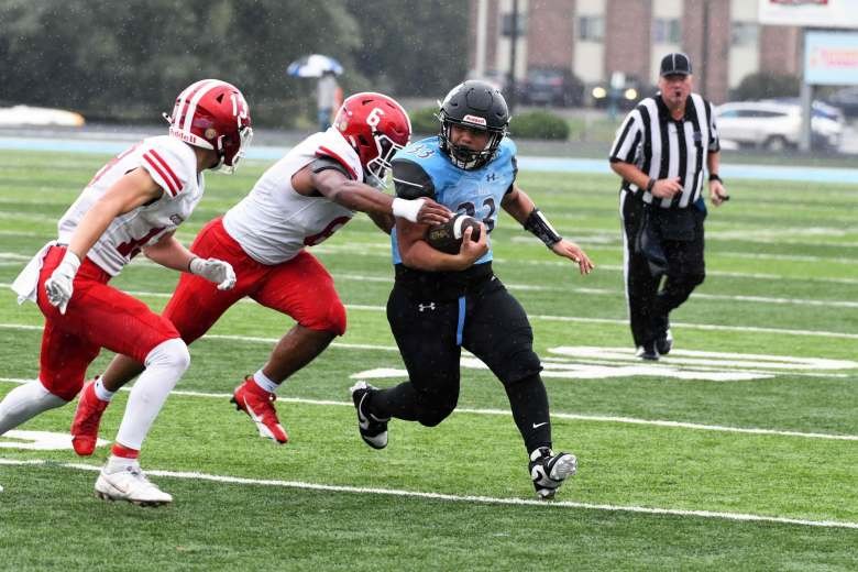 A ball carrier in a dark uniform stiff-arms a defender on a wet turf field.