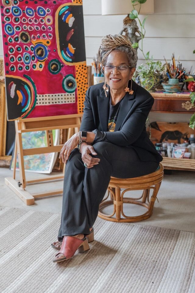 Woman with locs in a black blazer seated on a rattan stool before a colorful painting.
